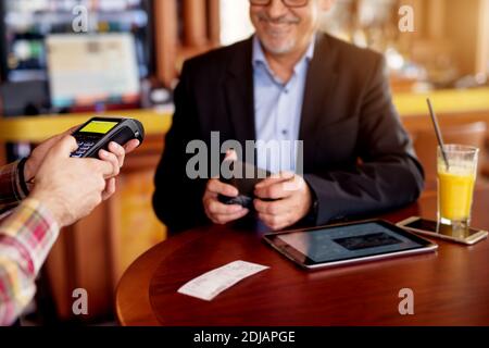 L'uomo d'affari allegro maturo sta usando un tablet e pagando il controllo con la carta di credito mentre si siede in una caffetteria. Foto Stock