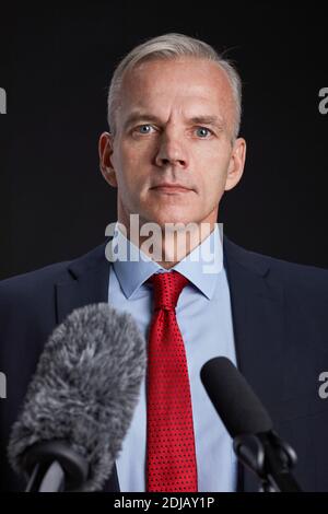 Ritratto verticale di uomo maturo in piedi sul podio e parlando al microfono su sfondo nero Foto Stock