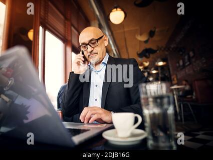 Un uomo d'affari professionale maturo è seduto al tavolo della caffetteria facendo una telefonata e utilizzando un computer portatile. Foto Stock