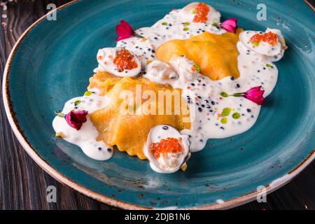 Ravioli con granchio in salsa caviale su grande piatto turchese. Il piatto si trova su un tavolo di legno ed è drappeggiato con un panno leggero. Foto Stock