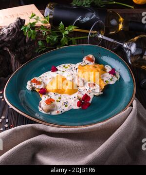 Ravioli con granchio in salsa caviale su grande piatto turchese. Il piatto si trova su un tavolo di legno ed è drappeggiato con un panno leggero. Foto Stock