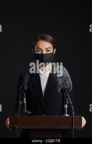 Ritratto verticale della giovane donna che indossa la maschera mentre dà la parola in piedi sul podio su sfondo nero Foto Stock
