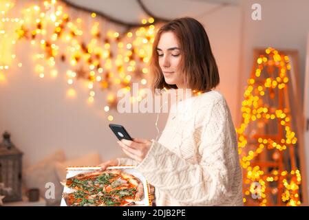 La giovane donna sta tenendo un telefono e una pizza a casa. Sta ordinando o scrivendo un feedback. Foto Stock