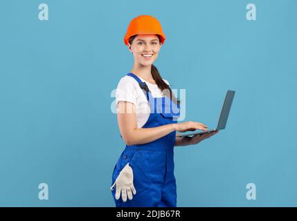 Donna in tute e elmetto utilizzando il computer portatile sopra sfondo blu Foto Stock