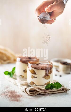 Tradizionale dessert italiano tiramisù in un bicchiere e la mano della donna spruzzare polvere di cacao su sfondo chiaro Foto Stock