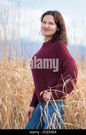 Bella giovane donna elegante vestita in pullover marrone, jeans blu in piedi e in posa in erba secca in campo in autunno Foto Stock