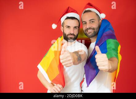 Felice coppia gay a santa Hat e avvolto con la bandiera omosessuale con pollici su sfondo rosso. Foto Stock
