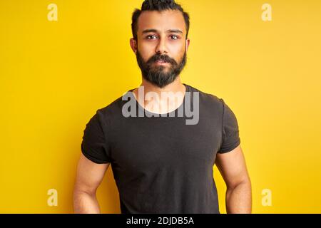 il maschio serio guarda con sicurezza la macchina fotografica, indossando una t-shirt nera casual. sfondo giallo isolato Foto Stock