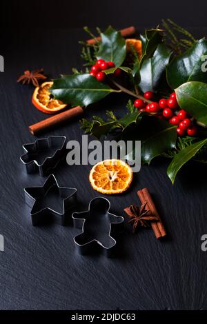 Concetto di fondo stagionale spezie invernali o natalizie, biscotti taglierina e Natale foglia agrifoglio e bacche su tavola nera in pietra ardesia con spazio per la copia Foto Stock