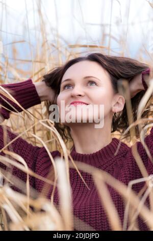 Ritratto di giovane donna elegante vestita di pullover marrone in posa in erba secca in campo. Donna felice con lunghi capelli ricci e labbra rosse. Foto Stock