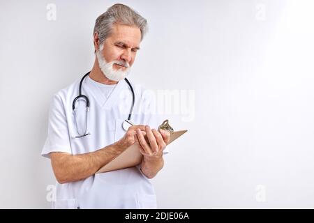 vecchio medico caucasico esperto che arita sintomi e diagnosticare, utilizzare la compressa per prendere appunti, indossare uniforme medica, isolato su sfondo bianco parete Foto Stock