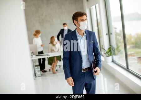 Bel giovane uomo d'affari che indossa una maschera facciale protettiva mentre si tiene tablet digitale in ufficio Foto Stock