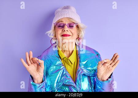 donna anziana in elegante indossare meditating isolato su sfondo viola, attraente donna mantiene la calma, in posa yoga Foto Stock