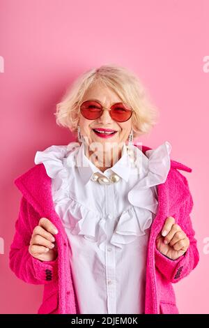 felice ottimista anziana signora in blusa e cappotto sorride alla macchina fotografica, indossando occhiali da sole, isolato su sfondo rosa Foto Stock
