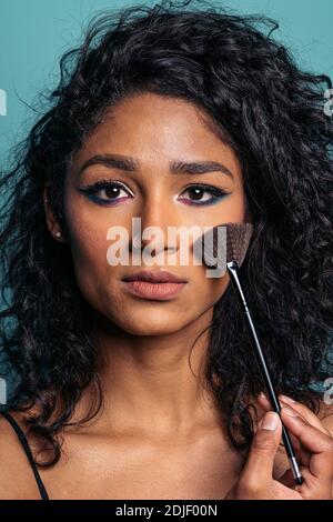 Foto di scorta di bella giovane donna ispanica che indossa il trucco colorato guardando la macchina fotografica e tenendo un pennello. Foto Stock