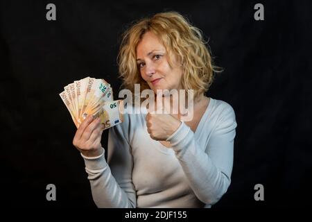Donna bionda di mezza età con un mazzo di banconote da 50 euro in una mano e il pollice dell'altra mano generato per indicare il successo Foto Stock