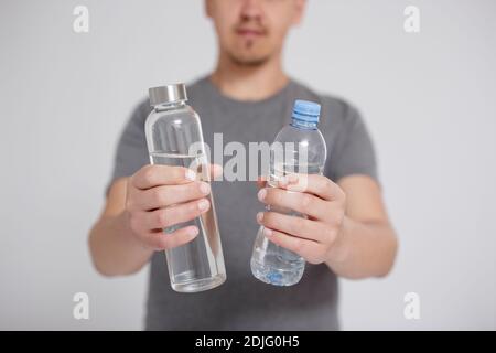 vetro o plastica, zero sprechi e concetto ecocompatibile - primo piano di acqua in vetro riutilizzabile e bottiglia di plastica a mani maschili su sfondo grigio Foto Stock