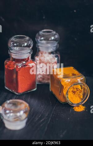 Un set di spezie in piccoli vasi con coperchi in sughero. Sale rosa, zenzero, curcuma, masala, paprika, paprika affumicata. Spezie su una superficie nera. Foto Stock