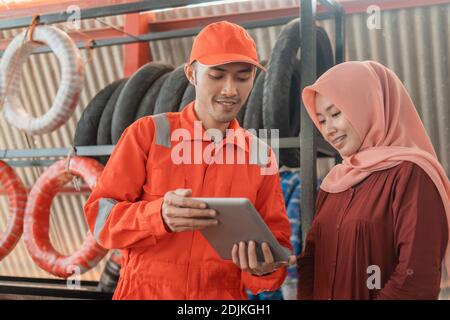 un meccanico maschile in un uniforme da indossare e una femmina cliente in un velo utilizzando un tablet digitale per la visualizzazione il catalogo presso un'officina di pezzi di ricambio Foto Stock