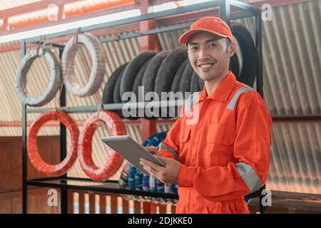 un mechanic sorridente in un dispositivo indossabile che utilizza un tablet digitale mentre si è all'interno l'officina con uno sfondo a cremagliera Foto Stock