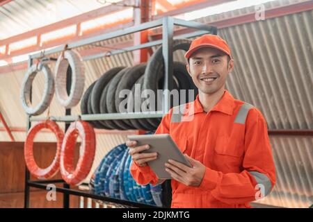 un meccanico asiatico sorridente in un dispositivo indossabile che utilizza un cuscinetto mentre si è all'interno l'officina con uno sfondo a cremagliera Foto Stock