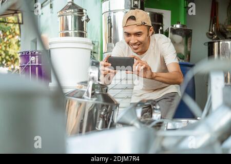 sorridente venditore maschile utilizzando la fotocamera del telefono quando si scatta una foto a. articoli per il negozio online presso il negozio di elettrodomestici Foto Stock