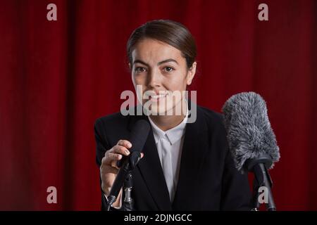 Ritratto di giovane donna che parla al microfono sul palco in piedi a podio contro la tenda rossa Foto Stock