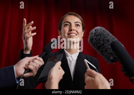 Vita in su ritratto di femmina sorridente ufficiale che dà intervista a. giornalista e parlando ai microfoni contro la tenda rossa Foto Stock