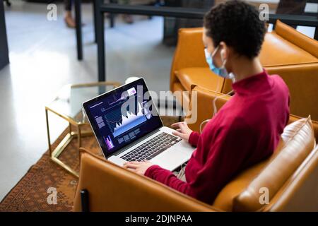 Donna mista che indossa maschera facciale che lavora su laptop indossando auricolari in ufficio casual Foto Stock