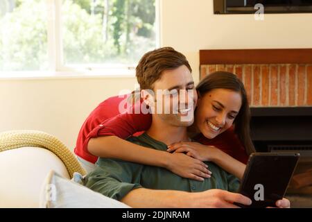 Coppia caucasica che ha videochiamata abbracciando e sorridendo al tablet in soggiorno Foto Stock