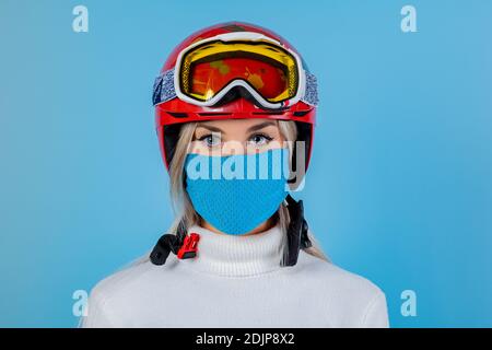 Primo piano ritratto di una ragazza sciatrice che indossa una maschera protettiva blu leggera, casco e occhiali. Uno snowboarder su uno sfondo blu chiaro. Covid19 coronavir Foto Stock