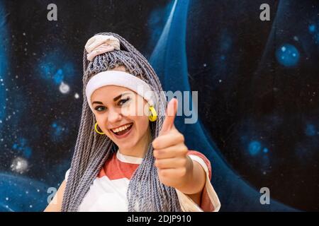 Ritratto di giovane donna eccitata con Dreadlock o kanekalon pigtail guardando la fotocamera e sorridendo mostrando pollici su indossando bianco fascia e yello Foto Stock