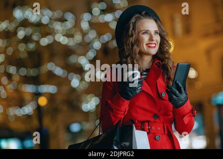 Winter Fun. Donna sorridente e alla moda con cappotto rosso e berta nera con borse per la spesa, tazza di latte di soia e smartphone che invia messaggi di testo all'esterno nel Foto Stock