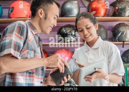 il cliente è in possesso di un portafoglio da pagare quando viene servito da un negoziante che tiene un tablet in un negozio di caschi Foto Stock