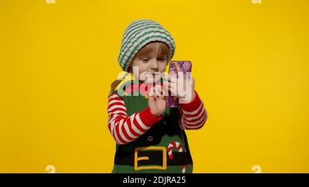 Vacanze di Capodanno. Gioioso bambino ragazza in costume elfo Natale aiutante Babbo fare una videochiamata sul cellulare a Babbo Natale o inviare messaggi di congratulazioni, regali online. Sfondo giallo. Foto Stock