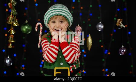 Impressed child kid girl in Christmas elf Santa Claus helper costume looks at camera makes big eyes covering mouth with hands feel horrified isolated on black background. OMG, wow, stunned, shocked Stock Photo