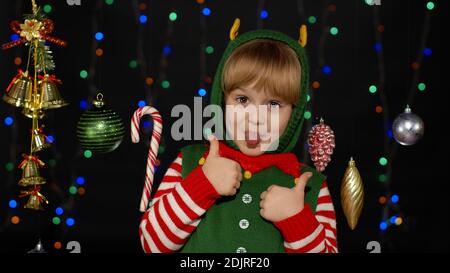 Teen kid girl in Christmas elf Santa Claus helper costume showing thumbs up gesture, agree sign on black background with garland decorations toys. Child smiling, showing tongue. New Year holidays Stock Photo