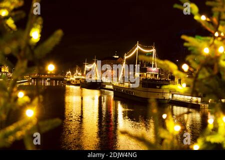 Barche da ristoranti ormeggiate lungo il fiume Aura con una splendida illuminazione notturna. Primo piano sfocato riempito con rami illuminati dell'albero di Natale. Foto Stock