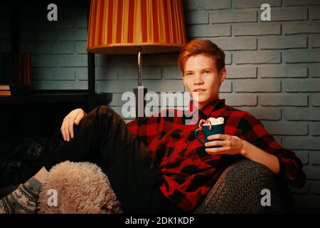 Bell'uomo in sedia regge tazza di cacao caldo con marshmallows. Drink di Natale con dolci e lecca. Parete in mattoni grigi e piantana su sfondo. Ragazzo in camicia plaid. Atmosfera festosa. Foto Stock