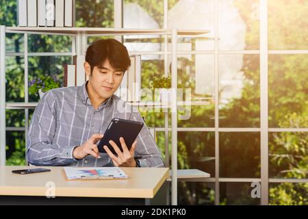 Giovane uomo d'affari asiatico seduto in ufficio sorride e lavora su un nuovo tablet, il giovane e sicuro che usa il touchpad checkin Foto Stock