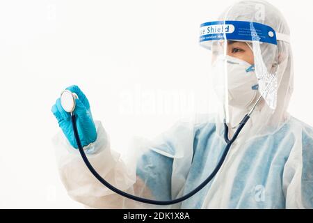 Closeup scienziato medico femminile in uniforme DPI che indossa un viso maschera protettiva e visiera protettiva in plastica che tiene sopra lo stetoscopio medico Mano per controllo Co Foto Stock