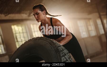 Donna forte che lavora fuori con un pneumatico enorme nello spazio di allenamento trasversale. Donna che si esercita con pneumatico grande in un magazzino abbandonato. Foto Stock