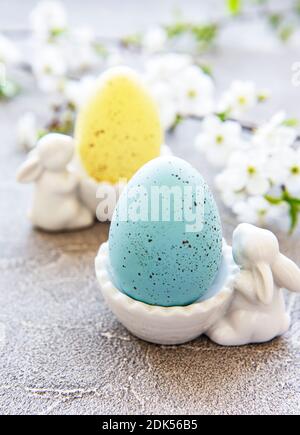 Uova di Pasqua colorate in piedi con figurine coniglietto di pasqua in ceramica e fioritura primaverile su fondo grigio in cemento Foto Stock