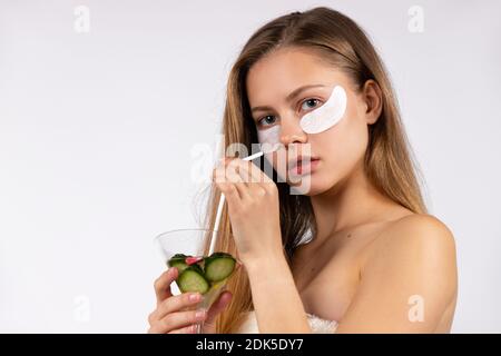 Biondo con macchie d'occhio sotto gli occhi, tenendo un cocktail detox cetriolo. Assistenza sana. Sfondo bianco. Foto Stock