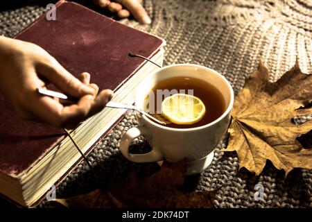 Accogliente foto delle mani femminili, una tazza di tè con limone sullo sfondo di maglia maglione, giallo foglie secche autunno e un vecchio libro, fuoco selettivo Foto Stock