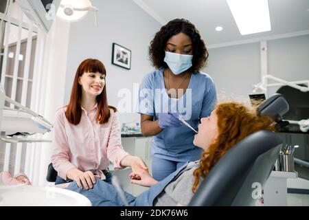 Trattamento dentale senza paura. Dentista africano femminile in uniforme blu e maschera, esaminando i denti della bambina, tenendo la mano di sua madre, a moderno Foto Stock