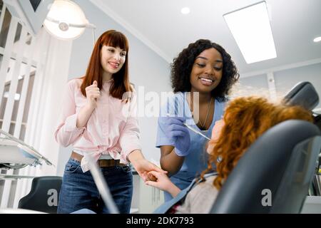 Mamma giovane donna che sostiene la sua figlia piccola presso l'ufficio del dentista durante il controllo. Donna sorridente pediatrica africana dentista in uniforme blu e guanti Foto Stock