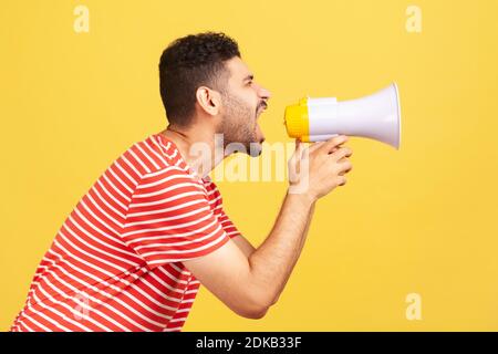 Profilo ritratto nervoso uomo bearded in t-shirt a righe a voce alta urlando tenendo megafono, annunciando messaggio importante. Riprese in studio al coperto Foto Stock