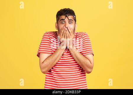 Scioccato uomo spaventato in t-shirt a righe che chiude la bocca con le mani, confuso e depresso con le informazioni, guardando con stupore e paura, la privacy. Foto Stock