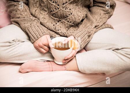 Una ragazza giovane donna in un maglione e calzini seduti a croce in un maglione sul letto e tiene una tazza con tè caldo e limone. Foto stile di vita Foto Stock